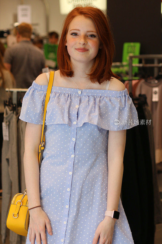 这是一个14 / 15岁的红发少女站在商店里的照片，她穿着一件漂亮的淡蓝色带纽扣的带有白色圆点图案的巴多连衣裙，肩上挎着一个黄色的手提包